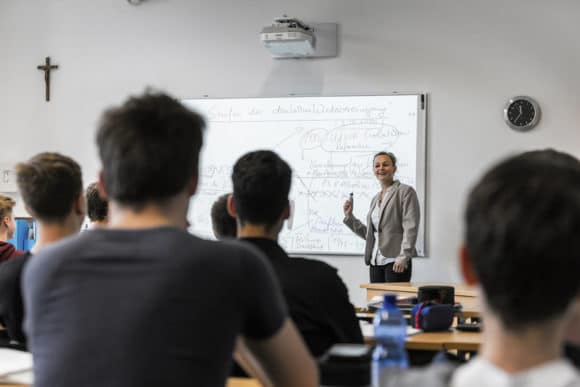 Klassenraum mit Lehrerin, Schüler:innen und Projektion