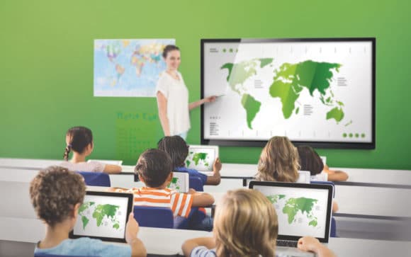 Klassenraum mit Lehrerin, Schüler:innen und interaktivem Großdisplay sowie Laptops und Tablets