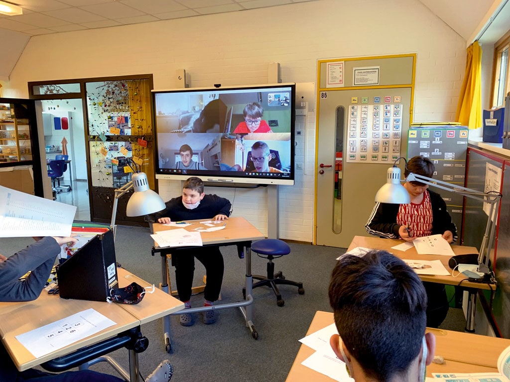 Klassenzimmer mit Kindern in Präsenz und über Webcam zugeschaltet auf Whiteboard