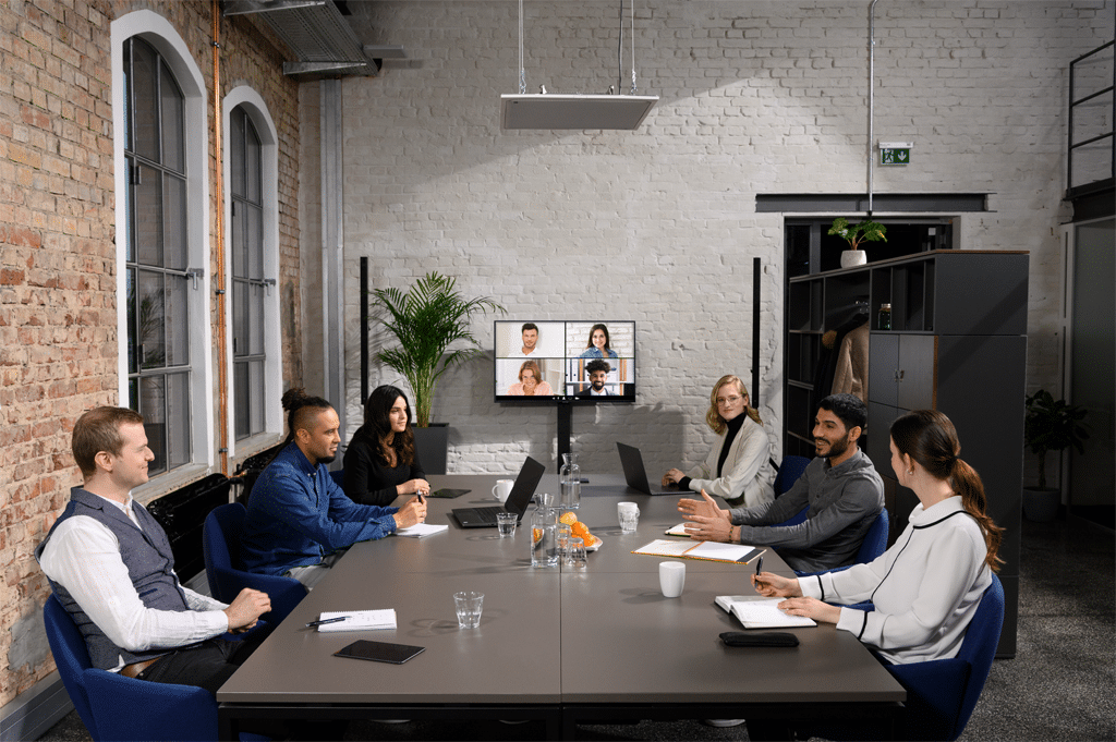 Drei Leute sitzen in einem Meetingraum, per Videokonferenz sind Leute dazugeschaltet