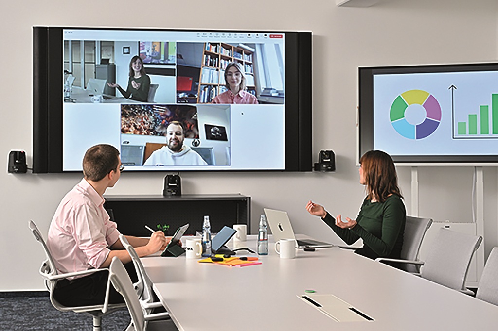 Frau und Mann an Konferenztisch in Videokonferenz mit großem Display an der Wand