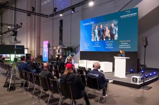 Zuhörer vor einem Podium, auf dem Jenny Köhler spricht, vor einem großen Display