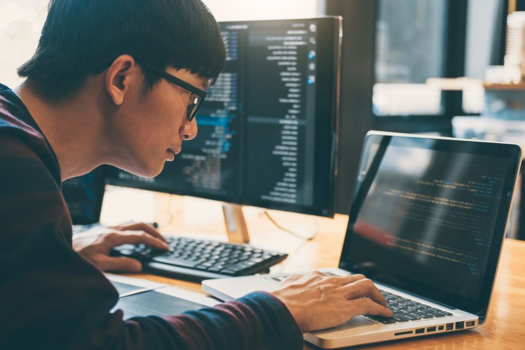 Softwareentwickler arbeitet an einem Laptop und einem zusätzlichen Monitor mit geöffneten Code-Ansichten.