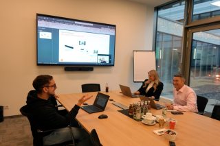 Drei Personen in einem Besprechungsraum mit großem Wanddisplay, Videobar und Laptops auf dem Konferenztisch.