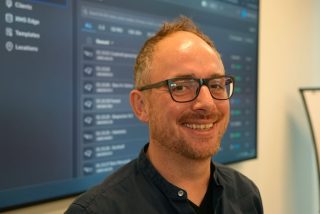 Porträt von Joachim Hopp vor einem Display mit der Benutzeroberfläche der zentralen Systemverwaltung.
