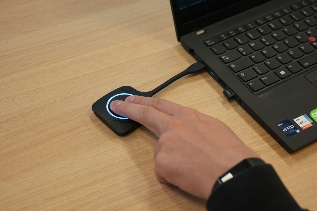 Nahaufnahme eines Barco ClickShare Buttons, der neben einem Laptop auf dem Konferenztisch bedient wird