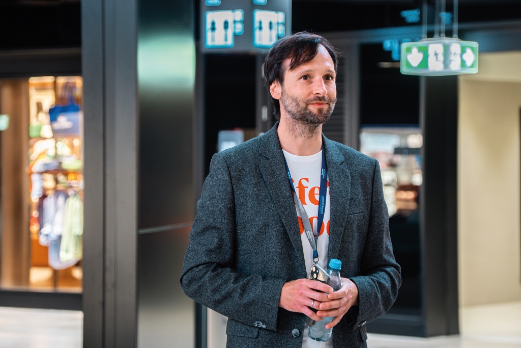 Mann mit dunklem Sakko und weißem T-Shirt steht mit einer Wasserflasche in der Hand im Terminal des Flughafens Frankfurt.