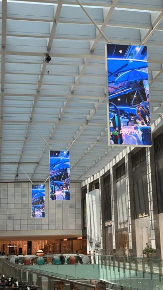 Drei hochformatige LED-Displays hängen hintereinander unter einem lichtdurchlässigen Deckenraster in der Dubai Mall, rechts Ladenfassaden über einer Galerie