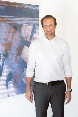 Jan Taube, CEO von Staige, steht vor einem großformatigen Bild an einer weißen Wand.
