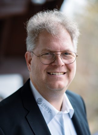 Porträt richard Werner. Lächelnder Mann mit grauem, lockigem Haar und Metallbrille in dunklem Sakko und weißem Hemd mit Trend-Micro-Logo am Kragen